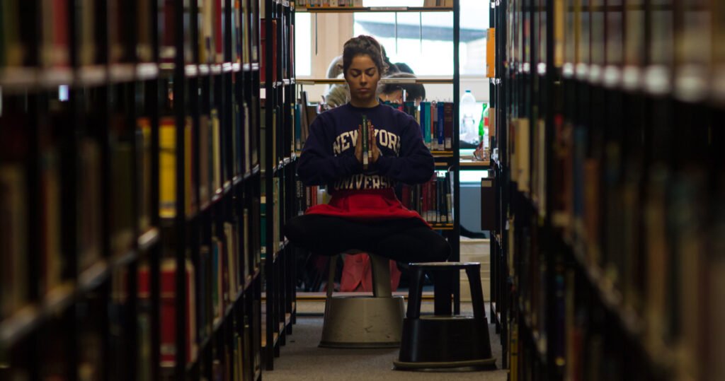 Carmen Mar focus position sitting on a chair in library holding 2 books in between hands anjali mudra